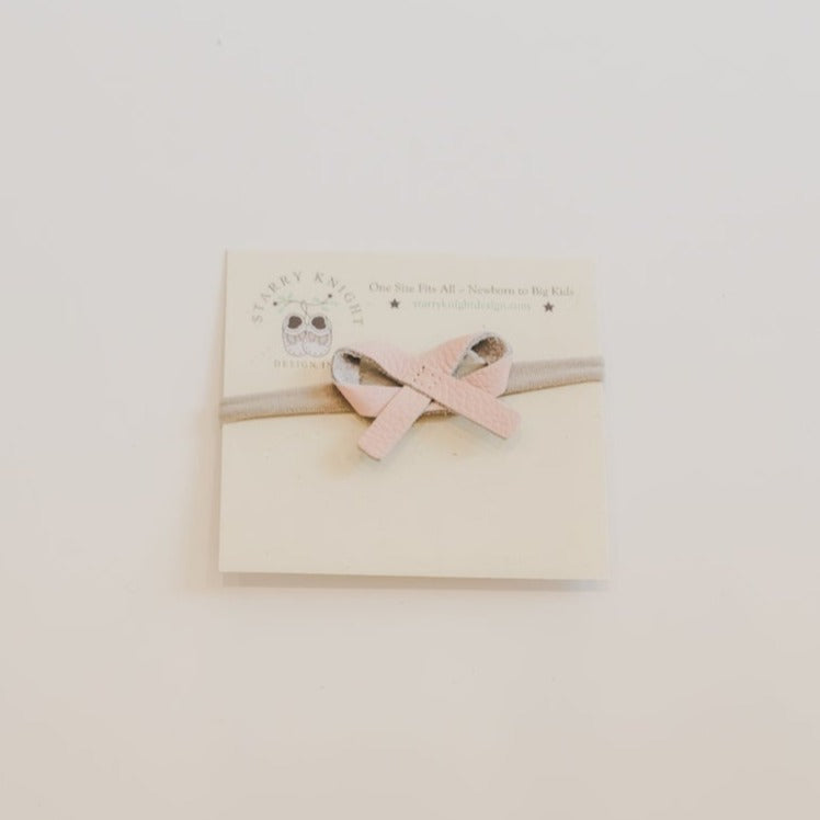 White background with the Ballet Bow Headband in Rose Blush by Starry Knight Design. Headband is on a nylon band, with a simple leather tie bow in a pink colour.