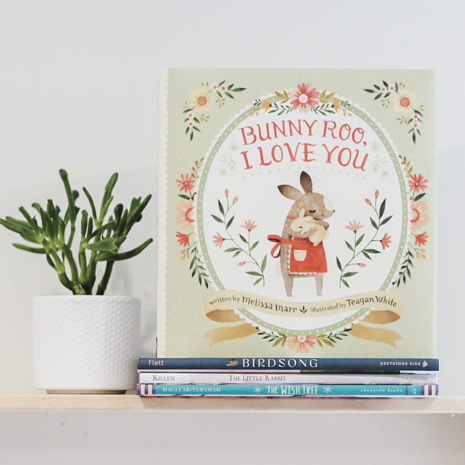 White wall with a wood shelf, a potted succulent and a stack of books with the book Bunny Too, I Love You by Melissa Marr on the top. Cover is green with a white oval in the middle and a mama & baby bunny hugging, and flowers all around.