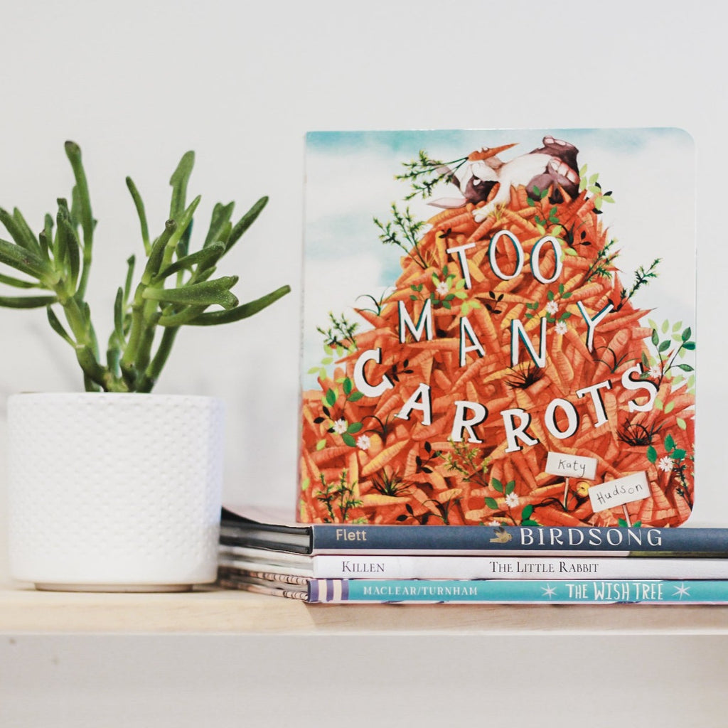 White wall with a wood shelf, a potted succulent and a stack of books with the book Too Many Carrots by Katy Hudson. Cover is a huge pile of carrots with a bunny laying on top holding one.