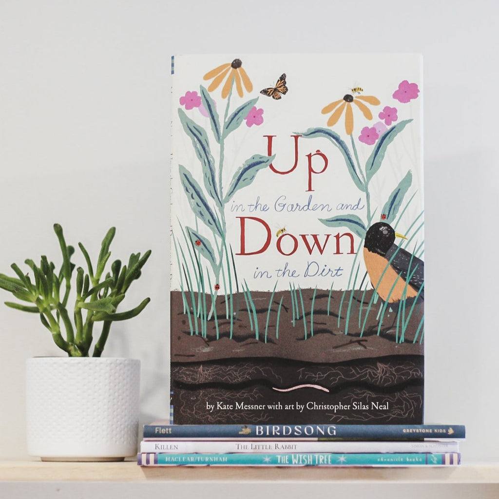 White wall with a wood shelf, a potted succulent and a stack of books with Up In The Garden and Down In The Dirt by Kate Messner. Cover is white with a close up of dirt and flowers growing, and a bird.