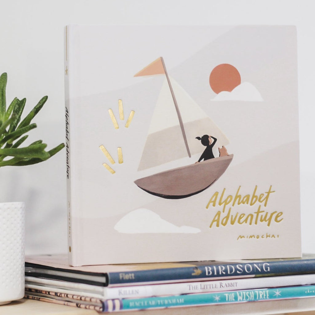 White wall with a natural wood shelf, a small succulent, and a stack of books with the book Alphabet Adventure by Mimochai on the top. The cover is a creamy grey colour, with a sail boat and it says Alphabet Adventure in gold embossed font.