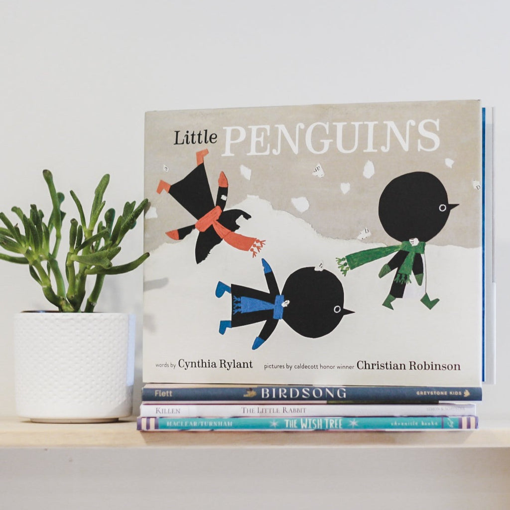 White wall with a wood shelf, a potted succulent, and a stack of books with the book Little Penguins by Cynthia Rylant on top. Cover is grey with some snow, and 3 penguins playing in the snow.