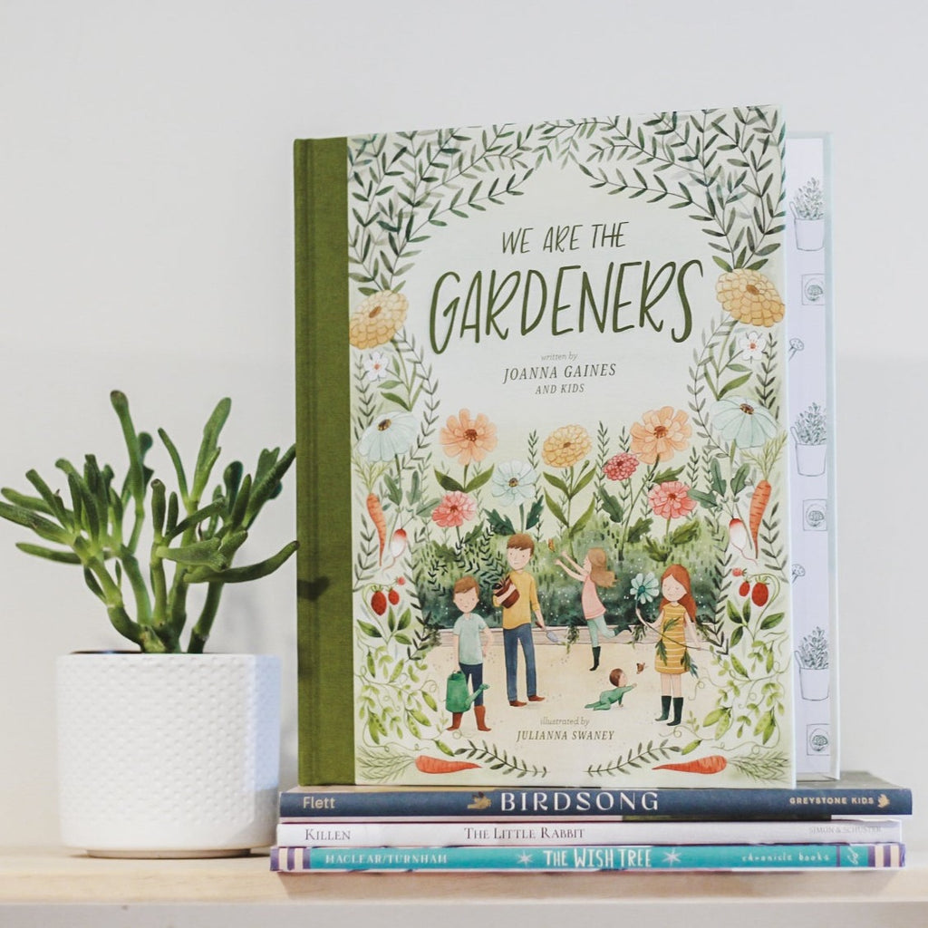 White wall with a wood shelf, a potted succulent and a stack of books with the book We Are The Gardeners by Joanna Gaines and Kids. Cover is light green with kids playing in a garden, and flowers all around.