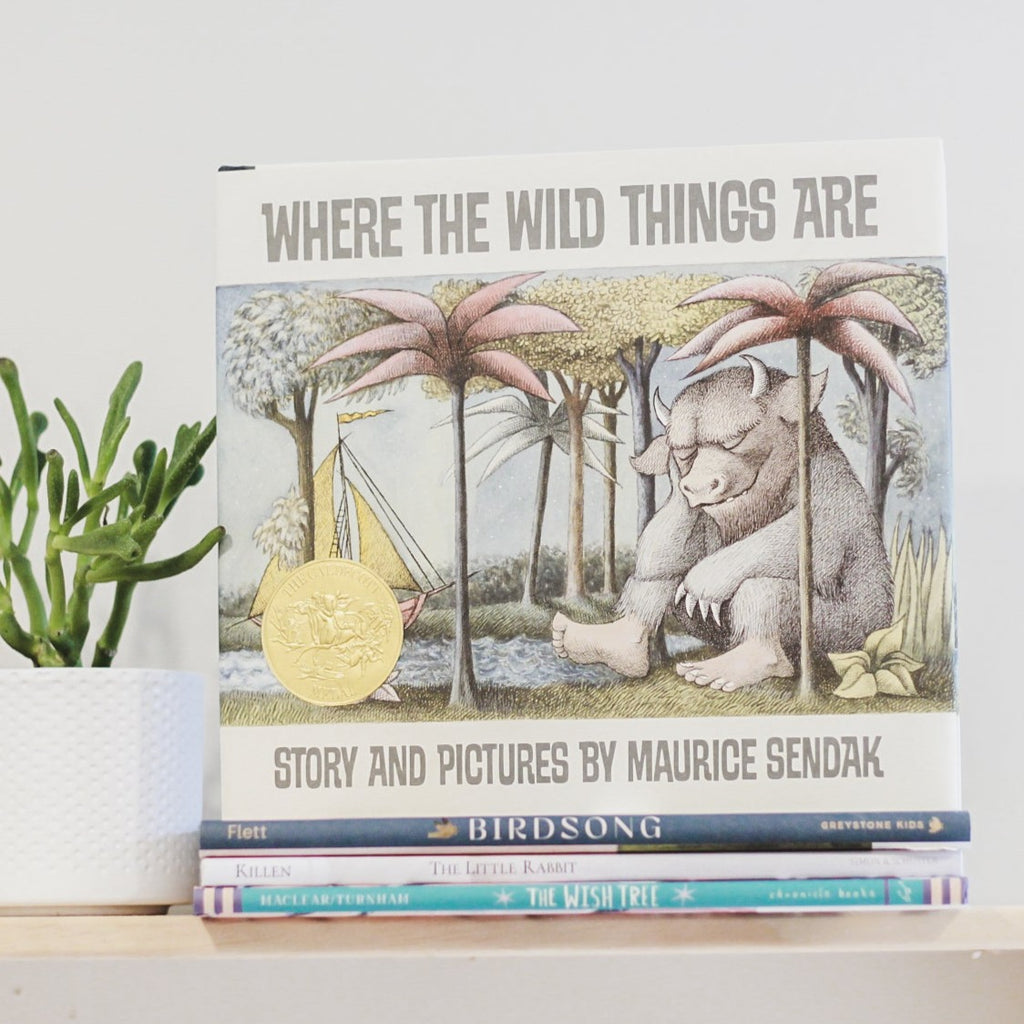 White wall with a wood shelf, a potted succulent and a stack of books with the book Where The Wild Things Are by Maurice Sendak. Cover is white with a wild thing sleeping under a tree.