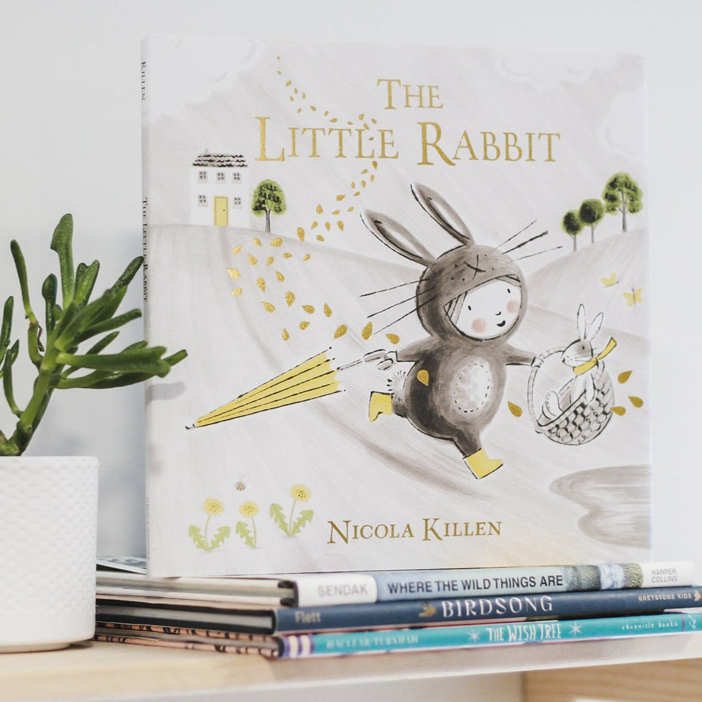 White wall with a wood shelf, a potted succulent, and a stack of books with the book The Little Rabbit by Nicole Killen on the top. The book is a neutral grey colour with a little girl dressed as a rabbit, running outside with an umbrella, and a basket with a bunny in it.
