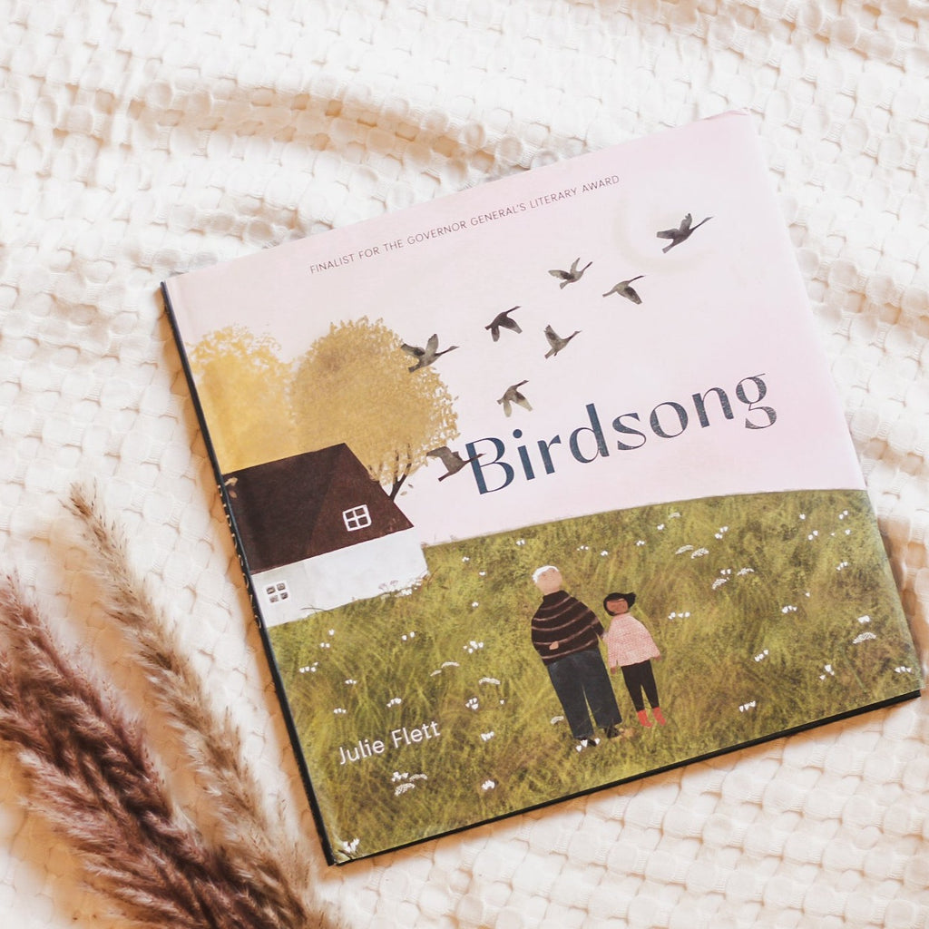 Waffle blanket background with the book Birdsong by Julie Fleet on top, and pampas grass to the side. Book cover is a pink sky with a White House, green grass, and a grandfather & girl standing outside, and birds in the sky.