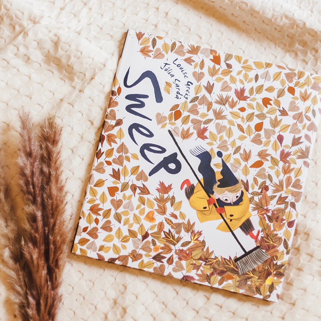 Cream waffle throw background with pampas grass in the bottom left corner and the book Sweep by Louise Greig. Cover is white with a boy pushing a broom throw autumn leaves.