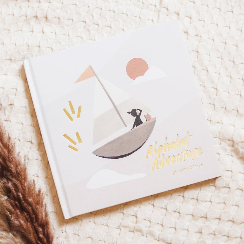 Cream waffle knit background with pampas grass on the bottom left corner, and the book Alphabet Adventure by Mimochair. The book is a creamy grey colour, with a sailboat, and gold embossed font that says "Alphabet Adventure".