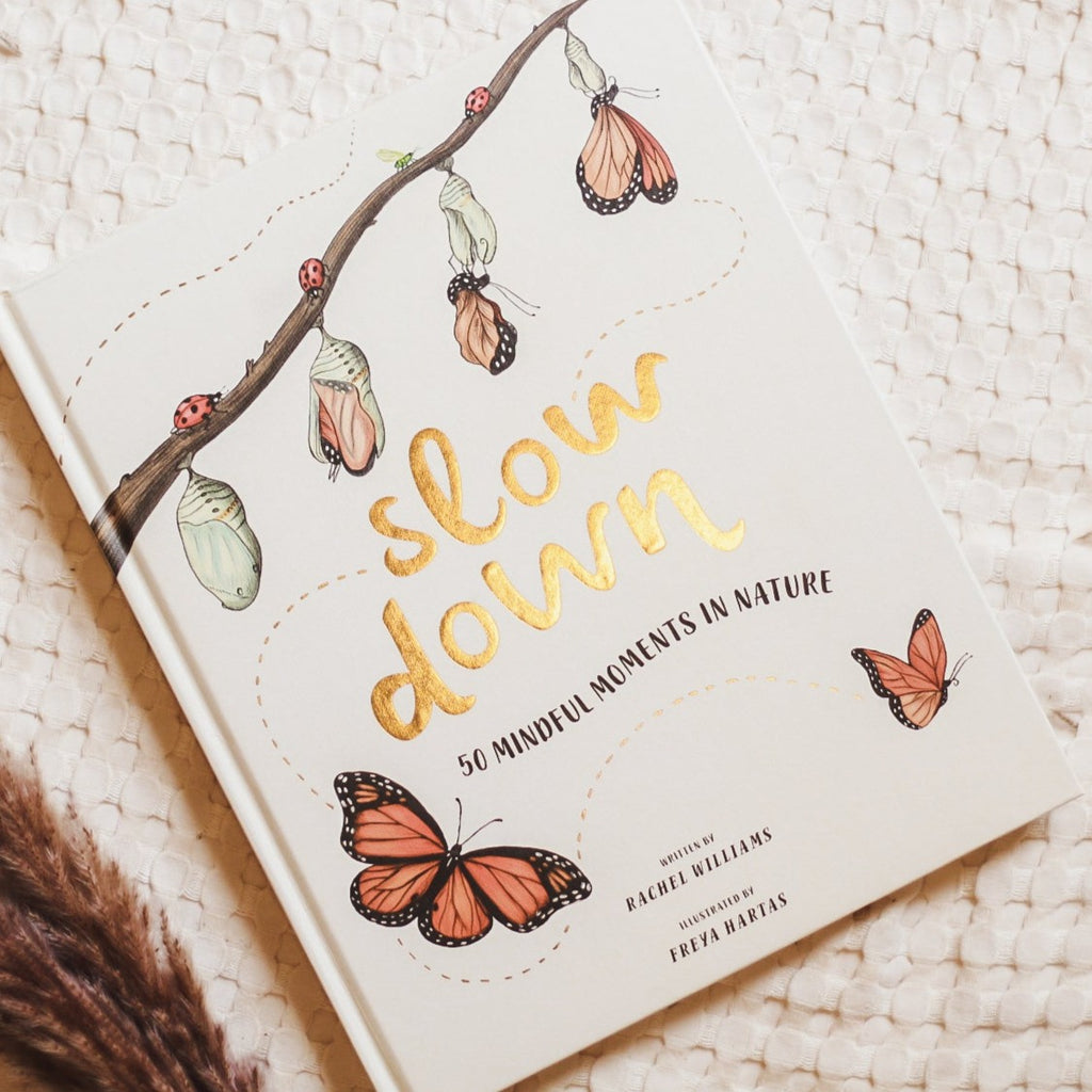 Cream waffle throw background with the book Slow Down: 50 Mindful Moments In Nature by Rachel Williams. Cover is a creamy white with the words "Slow Down" in gold embossed text, with the life cycle of a butterfly on top.