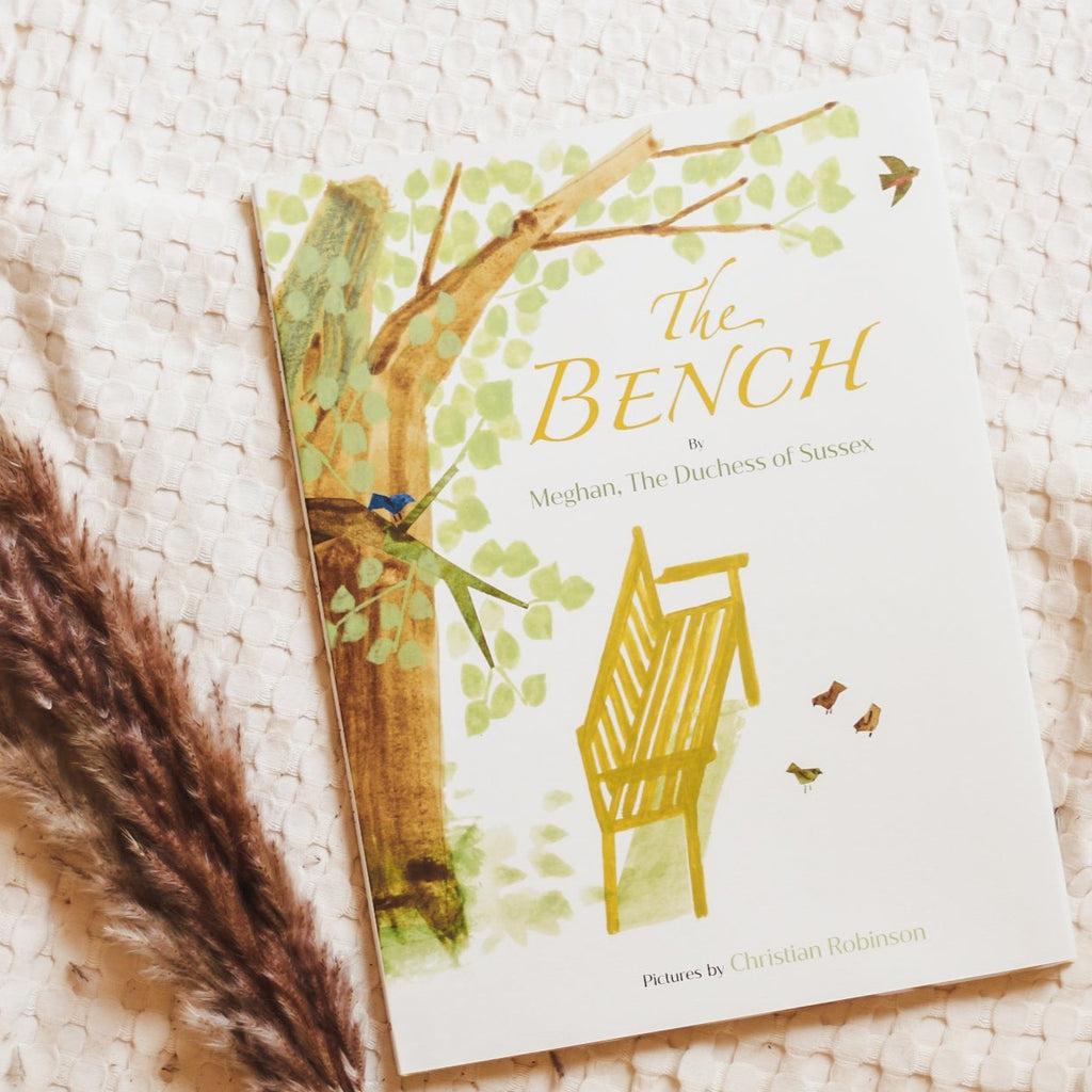 Cream waffle throw background with pampas grass on the bottom left side with the book The Bench by Meghan The Duchess of Sussex. Cover is cream with a wood bench, and a leafy green tree.