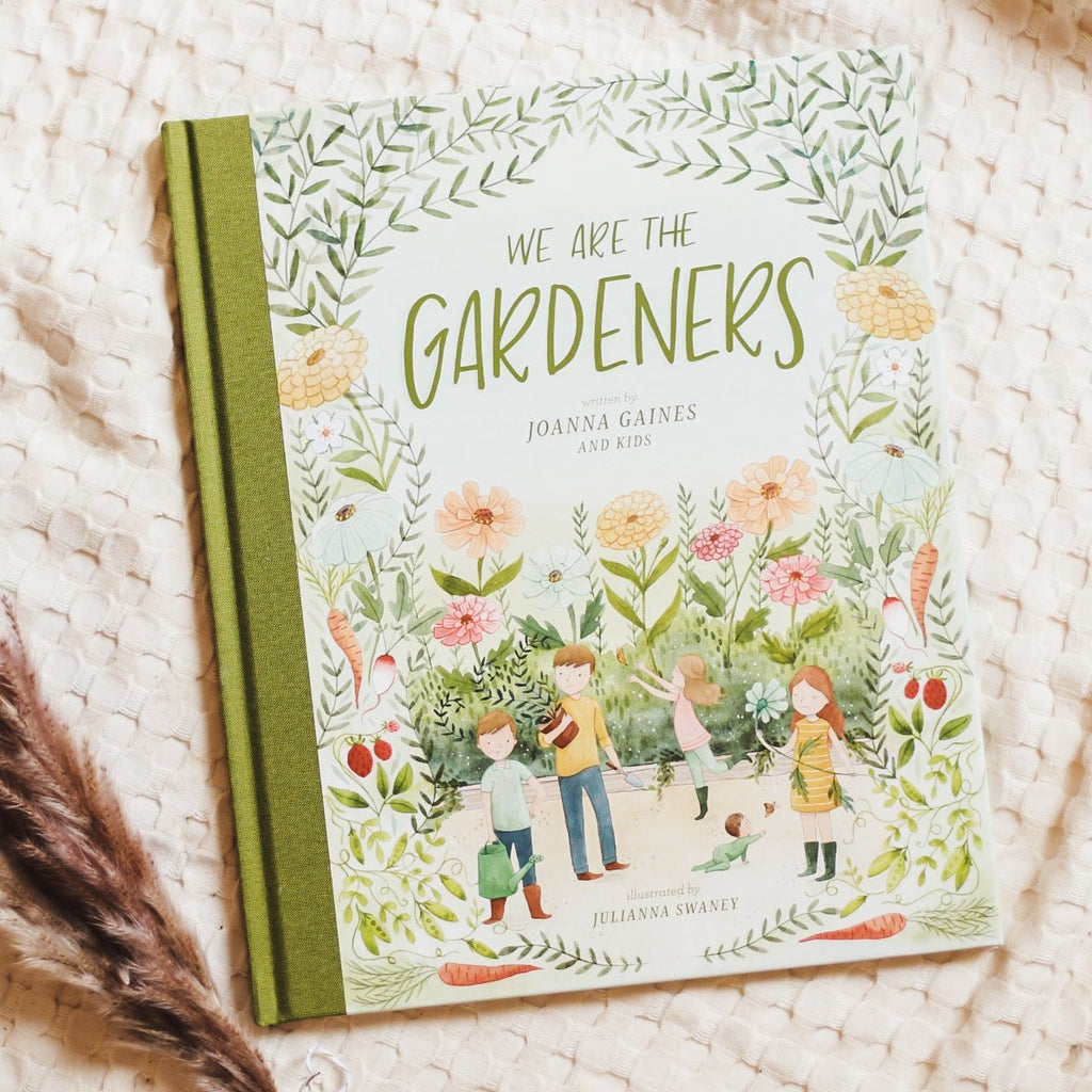 Cream waffle throw background with pampas grass on the bottom left side, and the book We Are The Gardeners by Joanna Gaines and Kids. Cover is green with kids playing in a garden, with flowers all around.