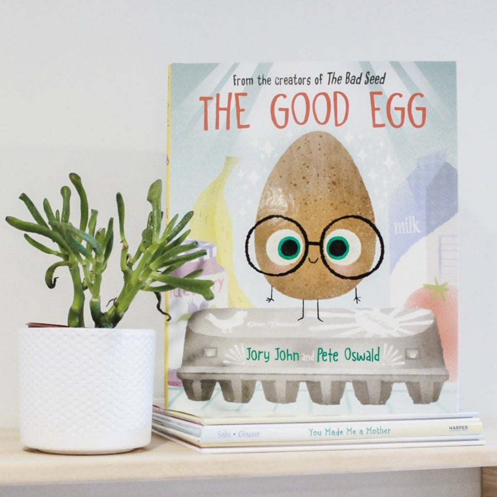 White wall with a natural wood shelf, and a succulent in a white planet, with the good The Good Egg by Jory John and Pete Oswald. Cover is an egg standing on a carton, with milk and bananas in the background.