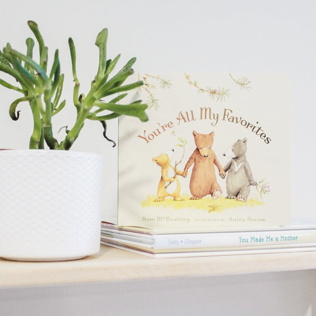 White wall with natural wood shelf, a succulent, a stack of books with the book You're All My Favorites by Sam McBratney on top. Cover is cream with a patch of grass with 3 bears holding hands.
