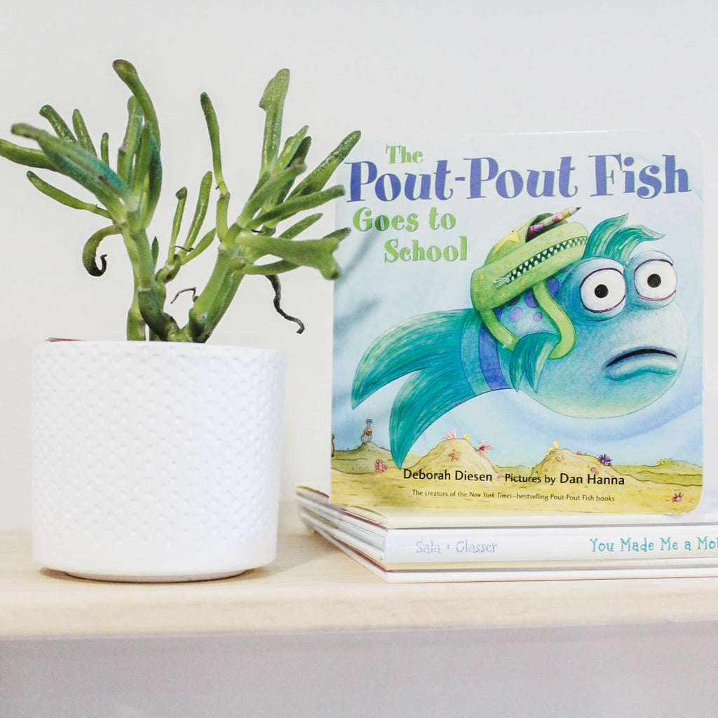 White background with a natural wood shelf, a green succulent on the left and The Pout-Pout Fish Goes To School book by Deborah Diesen. Cover is a fish swimming with a green backpack on.