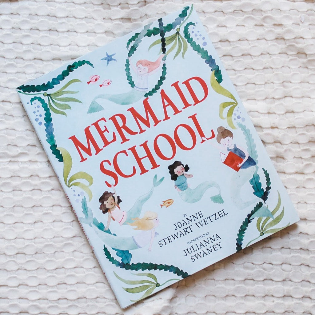 Overhead view of the book Mermaid School by Joanne Stewart Wetzel on a cream waffle knit blanket. Cover is blue with seaweed, and watercolour looking mermaids all over, and the title "Mermaid School" in bold red.