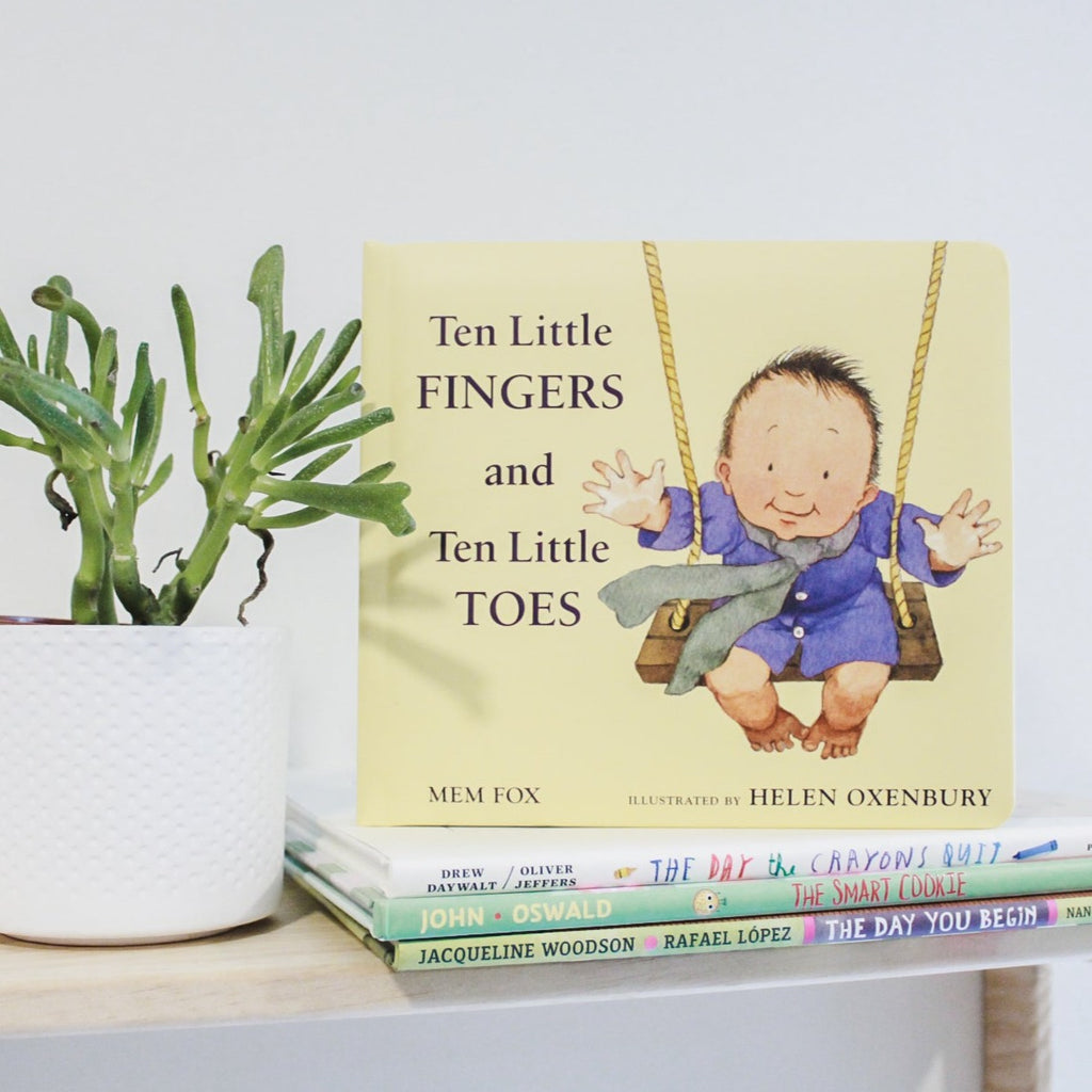 White wall with a natural wood shelf, a succulent, and a stack of books with the book Ten Little Fingers and Ten Little Toes by Mem Fox. The book is yellow with a drawing of a baby on a swing, reaching their arms out.