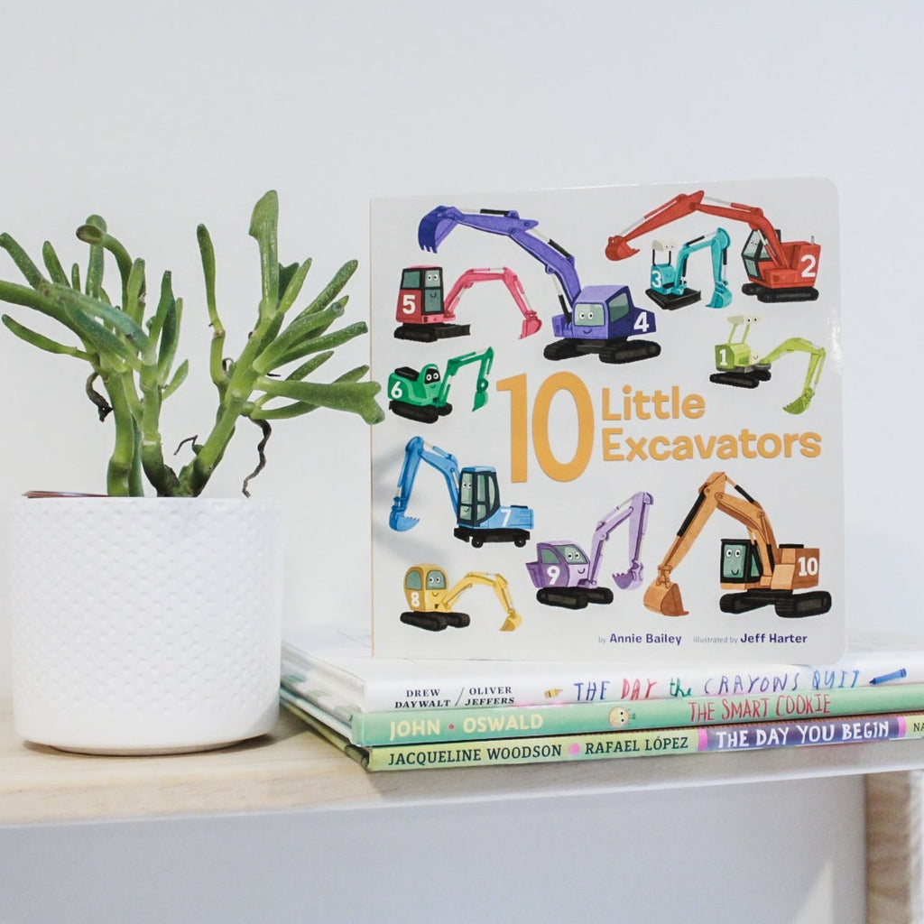 White wall with a natural wood shelf, a potted succulent, and a stack of books with the book 10 Little Excavator by Annie Bailey. White background with 10 Excavators in different colours all over.