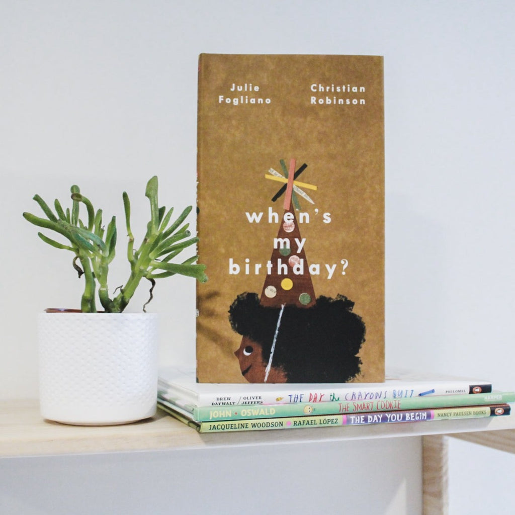 White background with a natural wood shelf, a succulent, and a stack of books with the book When's My Birthday? by Julie Fogliano on top. Book cover is a medium brown with a little girl looking to the side, wearing a party hat.