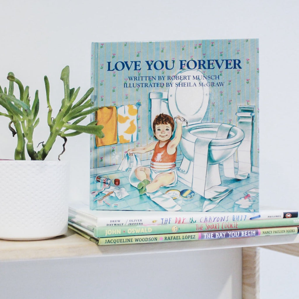 White background with a natural wood shelf, a green succulent, and a stack of books with the book Love You Forever by Robert Munsch on top. The book cover is blue with a baby sitting on the floor beside a toilet, with toilet paper, and a mess everywhere.