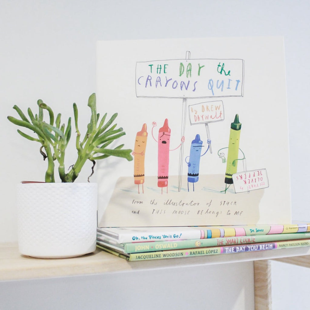 White background with a natural wood shelf, and a succulent, with a stack of books and the top one is The Day the Crayons Quit by Drew Daywalt. Cover is white with different coloured crayons holding up signs.