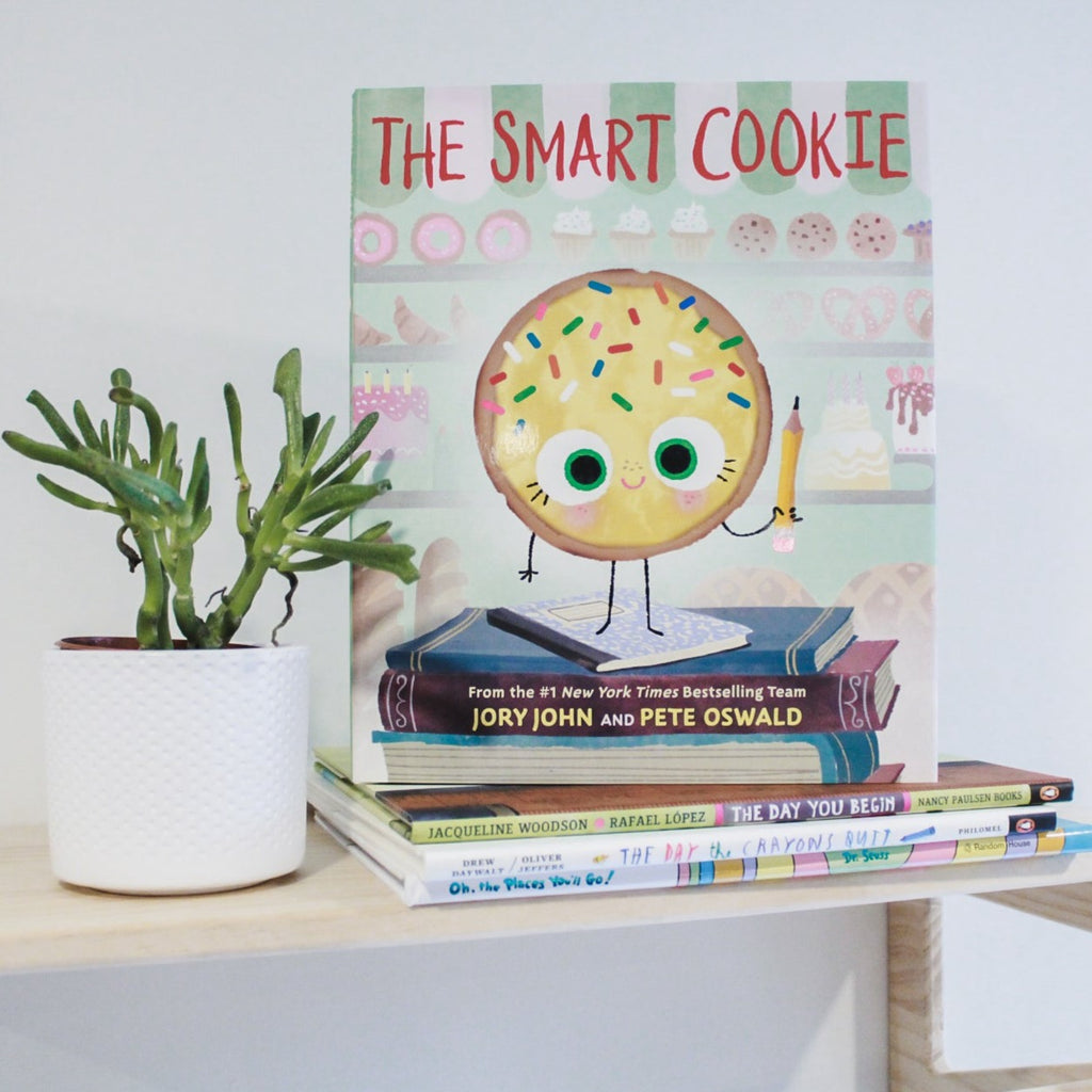 White wall with a natural wood shelf, a succulent on the left and a stack of books to the right of it, with the book The Smart Cookie by Jory John & Pete Oswald on top. The cover is a sugar cookie standing on top of a stack of books holding up a pencil, smiling.