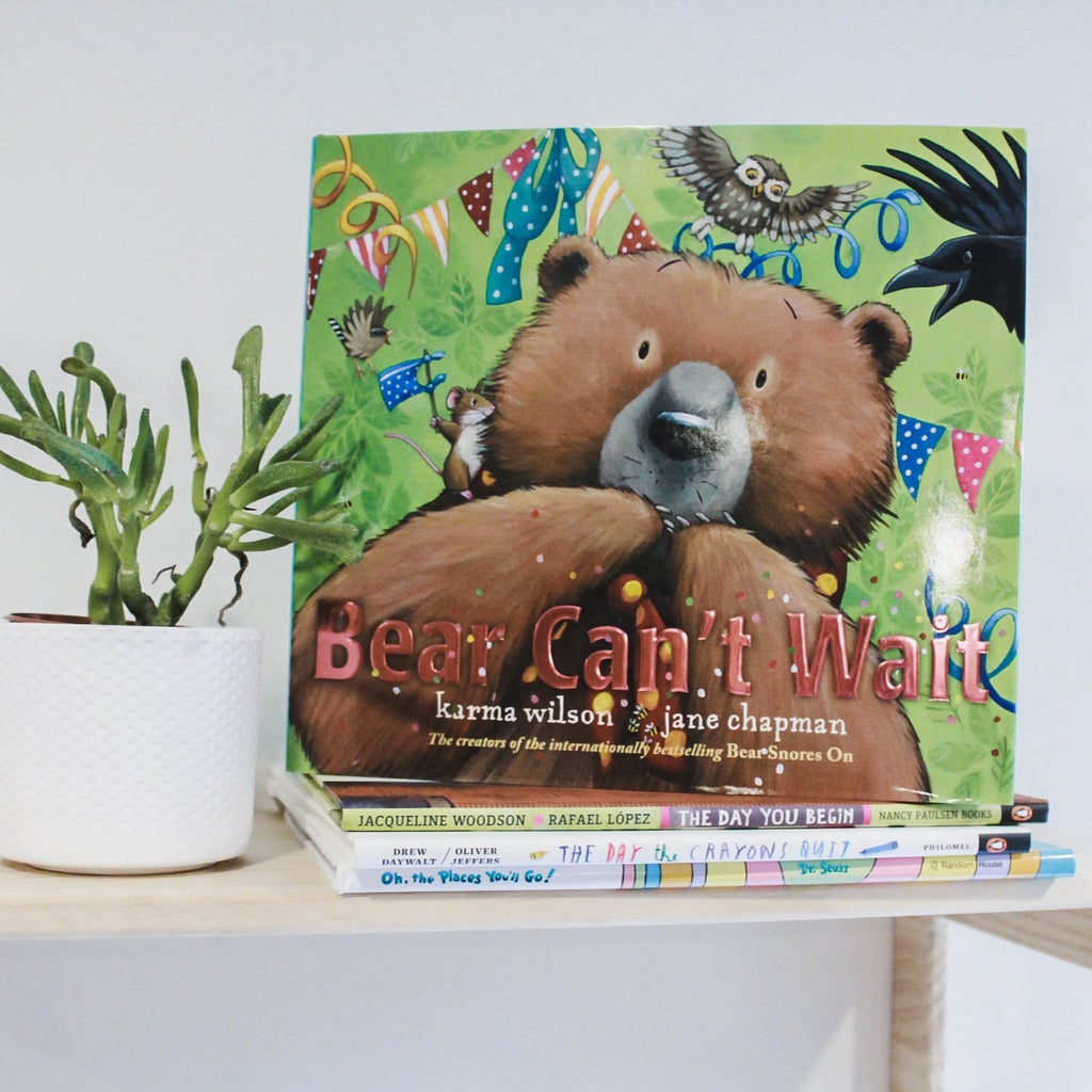 White background with natural wood shelf, a succulent, and a stack of books with the book Bear Can't Wait by Karma Wilson. Cover is green with colourful banners hanging, confetti, an owl, some birds, and a close up of a bear covering his mouth with his paws.
