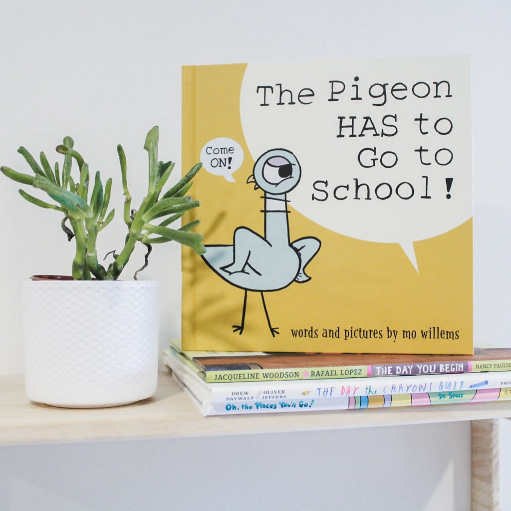 White wall with a natural wood shelf, a succulent, a stack of books, and the book The Pigeon HAS To Go To School! by Mo Willems. Cover is yellow with a pigeon on it, with a word bubble that says "Come ON!".