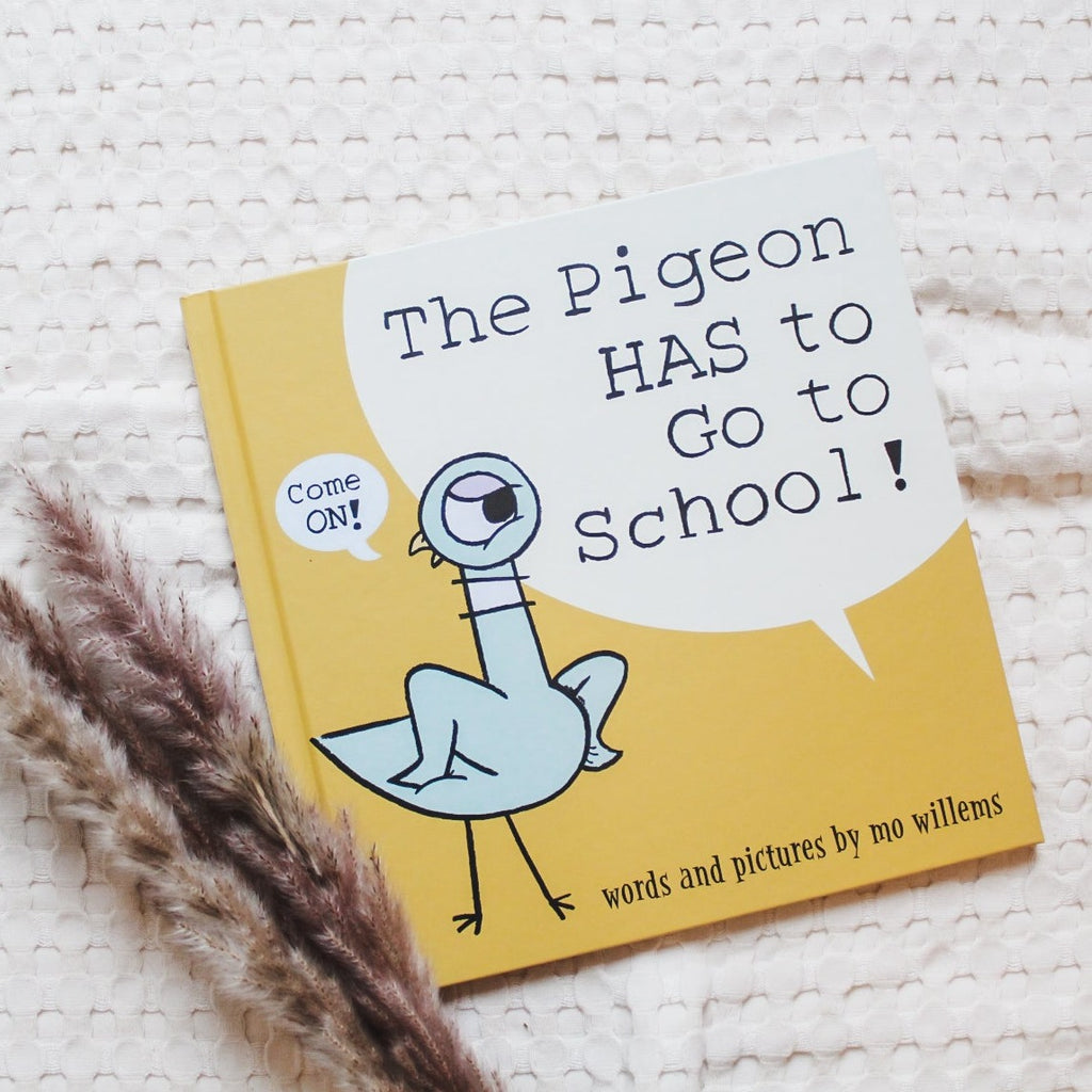 Overhead view of the book The Pigeon HAS To Go To SchooL! by Mo Willems, on a cream waffle knit quilt. Cover is yellow with a blue pigeon, and the words "Come On!" in a word bubble.
