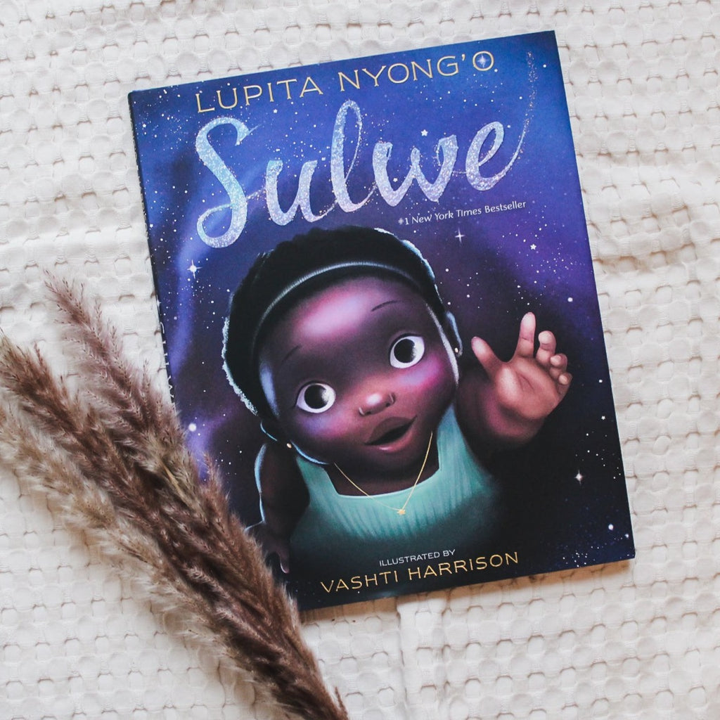 Cream waffle quilt background with pampas grass on the bottom left corner, and the book Sulwe by Lupita Nyong'o. Cover is purple, blue, and black.. with stars, like a night sky, and a little girl reaching her hand out.