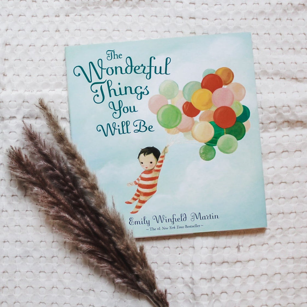 Cream waffle blanket background with pampas grass on the left corner, and the book The Wonderful Things You Will Be by Emily Winfield Martin. The cover is a blue sky with a baby holding floating in the air holding onto balloons.