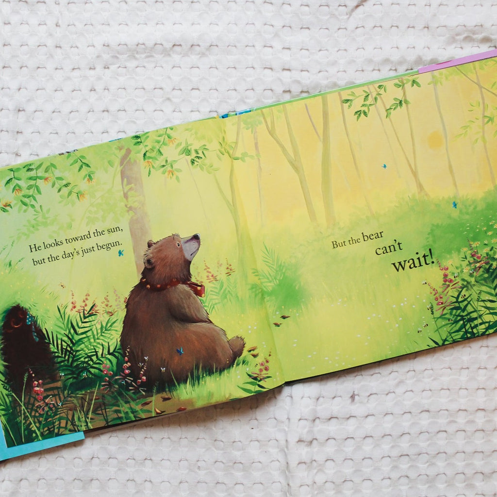 Overhead view of a page from Bear Can't Wait by Karma Wilson. Page has bear sitting in the grass looking at the sun rising, and the words say "He looks toward the sun, but the day's just begun. But the bear can't wait!"