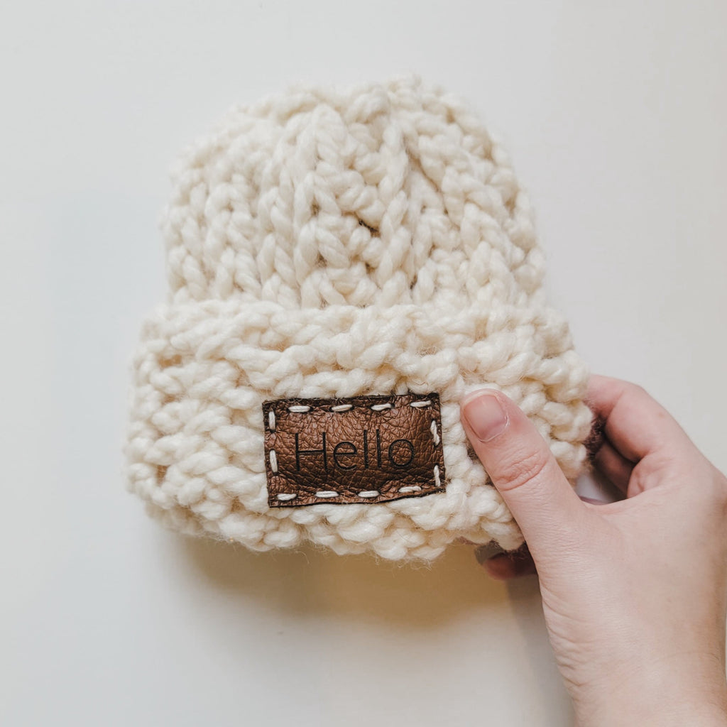 White background with Handknit "Take Me Home" Beanie in Natural by Petit Nordique, with a hand holding the side. Beanie is a cream colour with a sewn on leather patch on the front that says “Hello”.