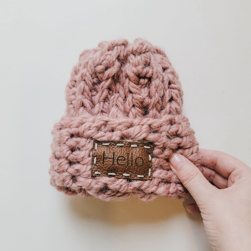 White background with Handknit "Take Me Home" Beanie in Rose by Petit Nordique, with a hand holding the side. Beanie is a rose colour with a sewn on leather patch on the front that says “Hello”.