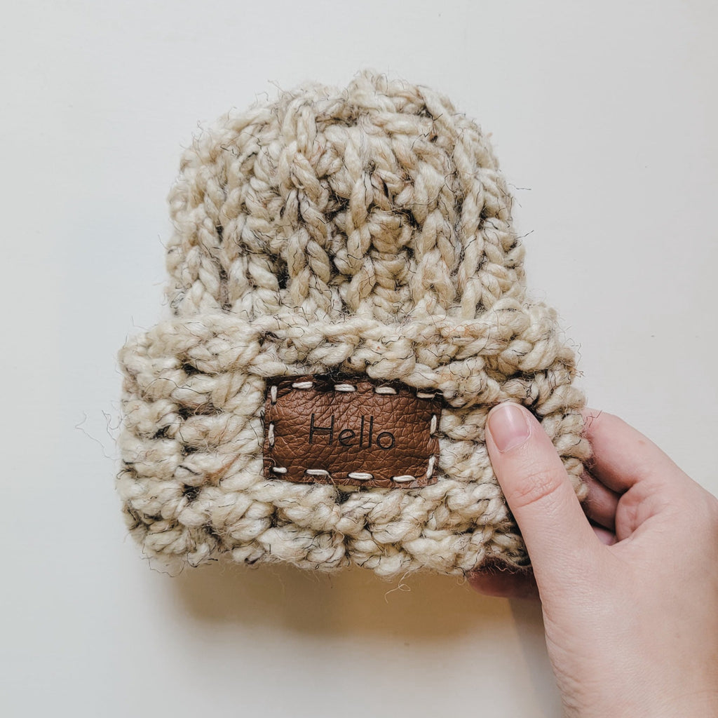 White background with Handknit "Take Me Home" Beanie in Oatmeal by Petit Nordique, with a hand holding the side. Beanie is an oatmeal tweed with a sewn on leather patch on the front that says “Hello”.