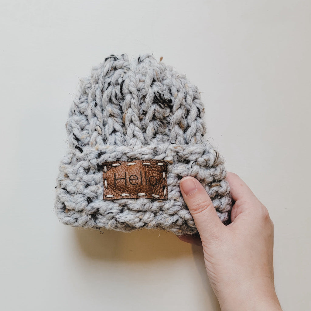 White background with Handknit "Take Me Home" Beanie in Grey Tweed by Petit Nordique, with a hand holding the side. Beanie is a grey tweed with a sewn on leather patch on the front that says "Hello".