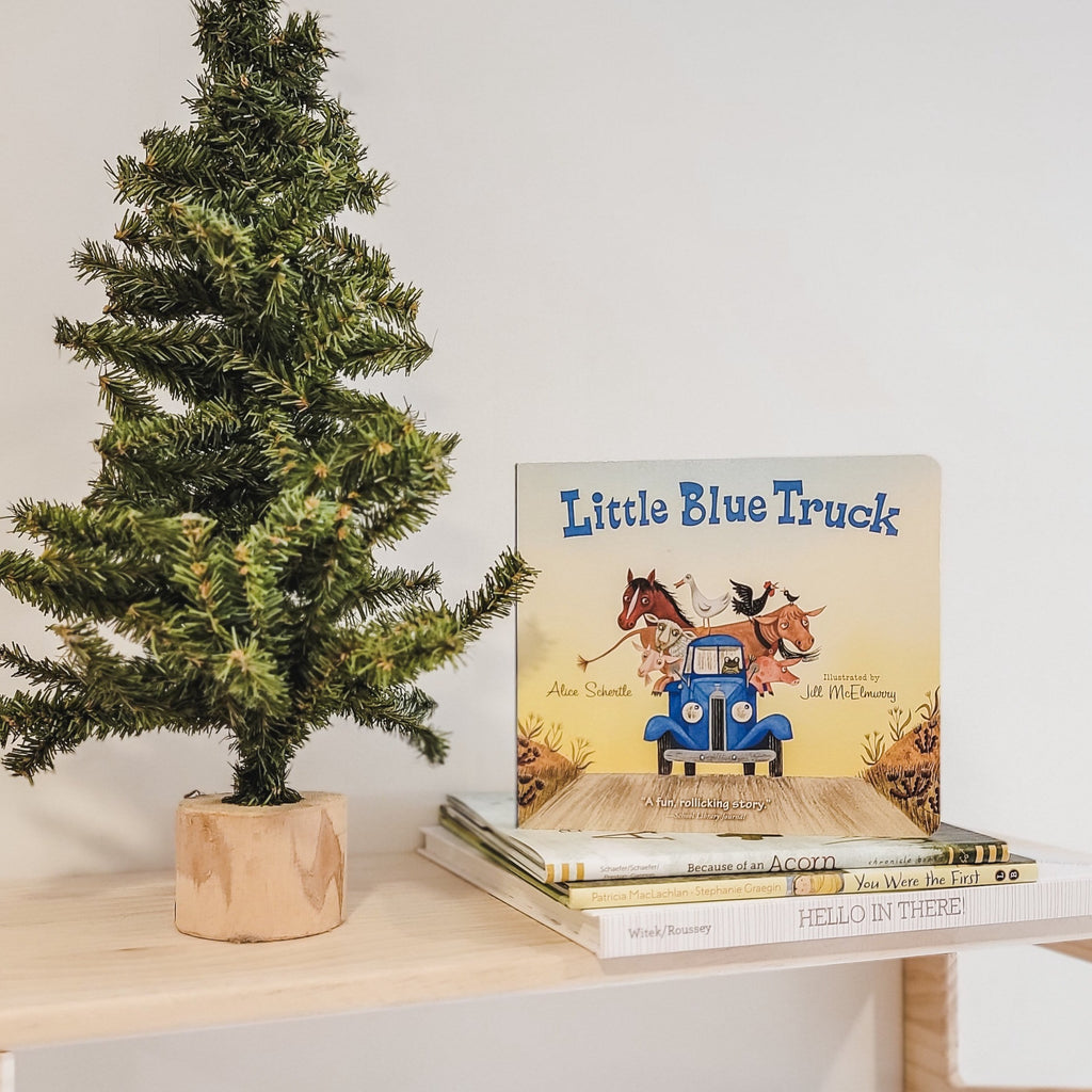 White background with a natural wood shelf with a small pine tree beside a stack of books, with the book The Little Blue Truck by Alice Schertle. Cover is the little blue truck with animals in the back of it.