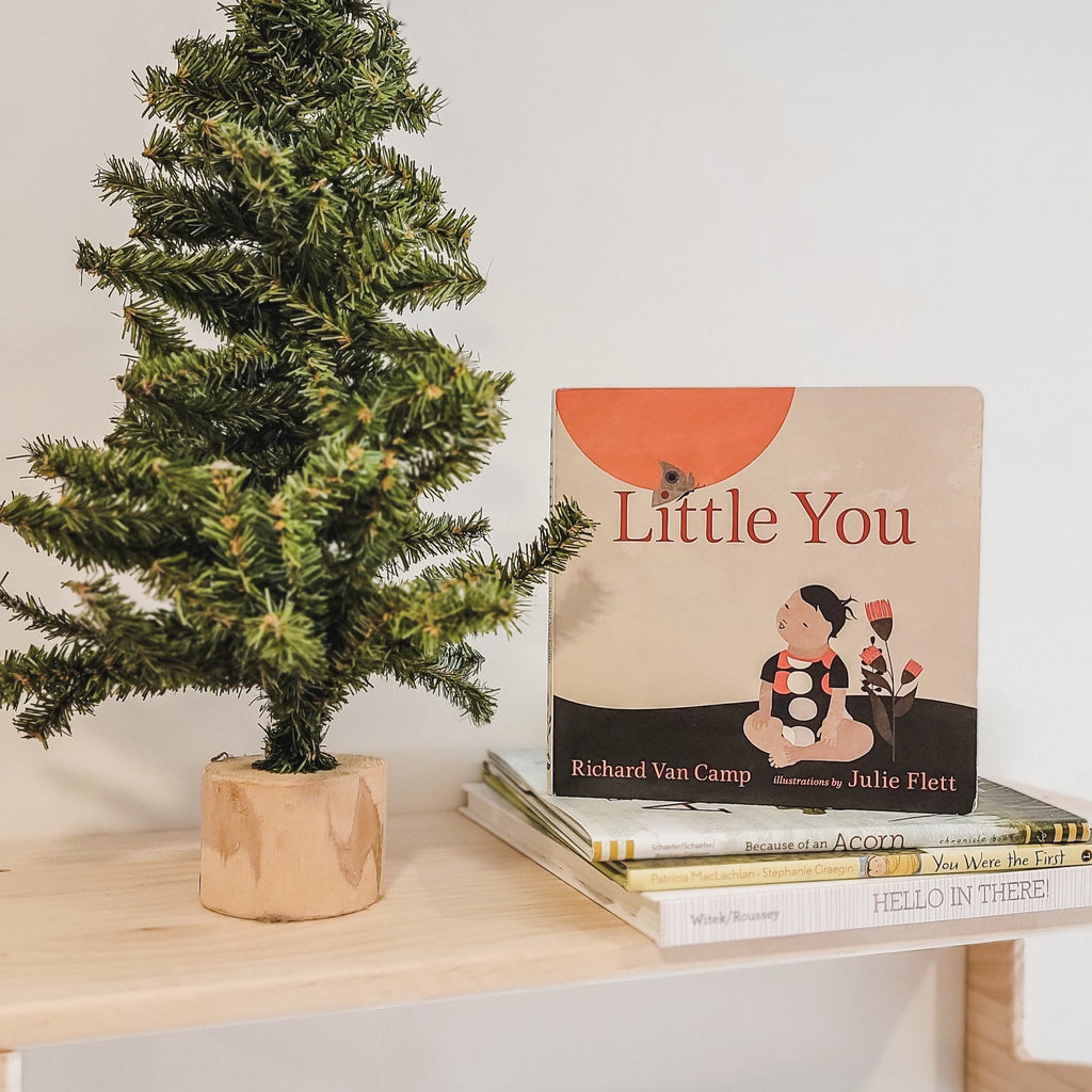 White wall with a natural wood shelf, and a small pine tree beside a stack of books with the book Little You by Richard Van Camp on the top. The book cover is beige with a black ground and a little person sitting down cross legged, with a flower beside them.