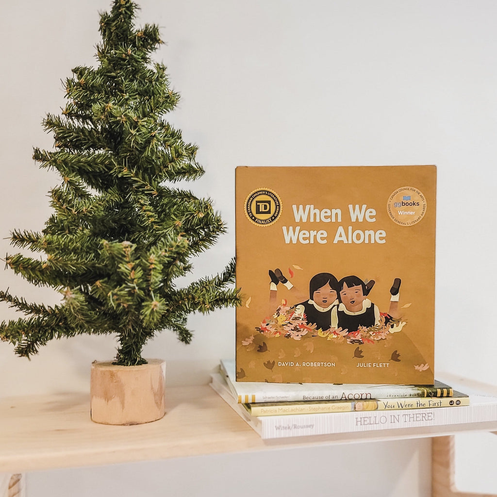 White wall with a natural wood shelf, and a pine tree beside a stack of books, the book When We Were Alone by David A. Robertson is on top. Cover is a brownish colour with 2 girls playing in leaves.