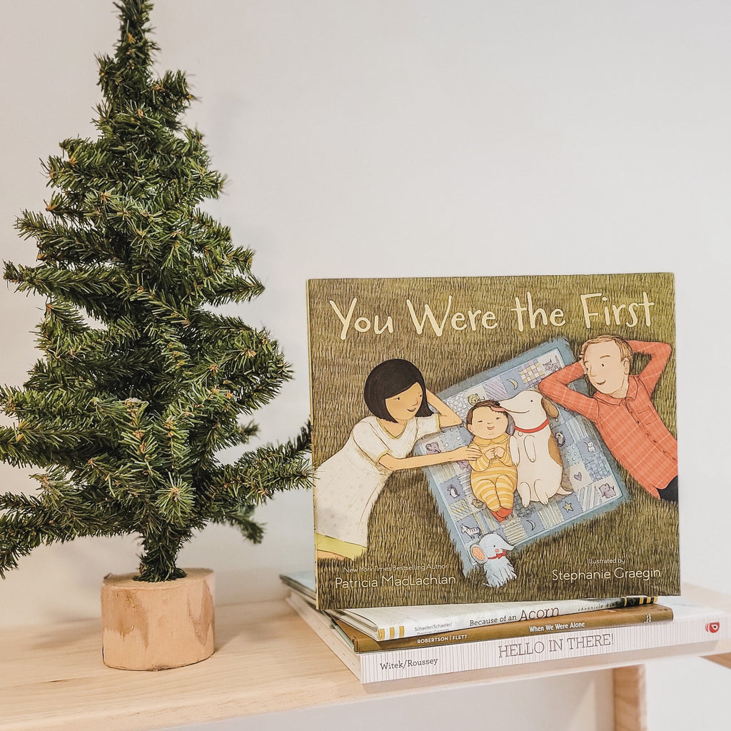 White wall with a natural wood shelf, a pine tree beside a stack of books with the book You Were The First by Patricia MacLachlan on top. Cover is a family laying in the grass together, baby on a blanket with their dog.