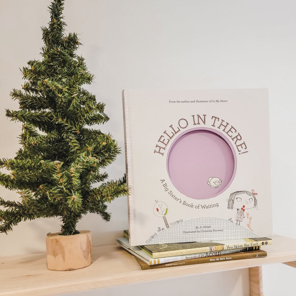 White wall with a natural wood shelf with a small pine tree beside a stack of books and the book Hello In There: A Big Sister's Book Of Waiting by Jo Witek on top. Cover is white with a big purple circle and a little girl standing on top of a hill