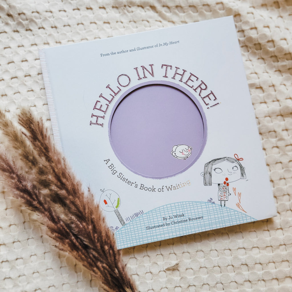 Cream waffle blanket with pampas grass on the bottom left corner and the book Hello In There: A Big Sister's Book Of Waiting by Jo Witek. Cover is white with a big purple circle with a baby in it, and a little girl standing on a hill with flowers.