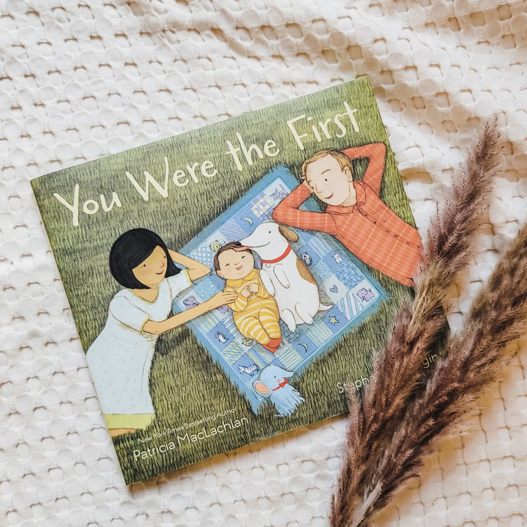 Cream waffle blanket with pampas grass on the bottom right corner, and the book You Were The First by Patricia MacLachlan. Book cover is a family laying in the grass, baby and dog on a blue blanket in the middle.