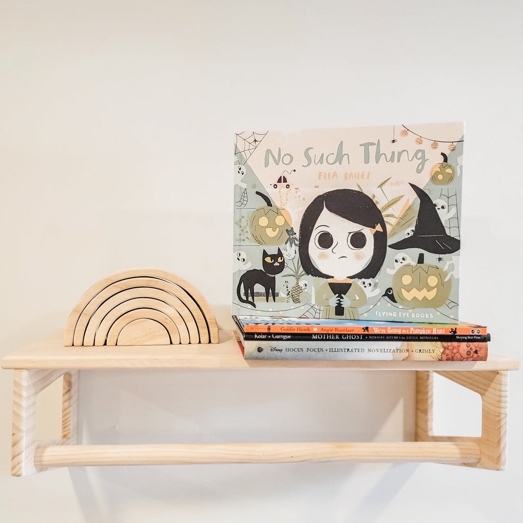 White background with a natural wood shelf with a wood rainbow stacker, and a stack of books with No Such Thing by Ella Bailey on top. Book is a blue colour with a little girl holding a flashlight, with pumpkins, ghosts, and cats all around.