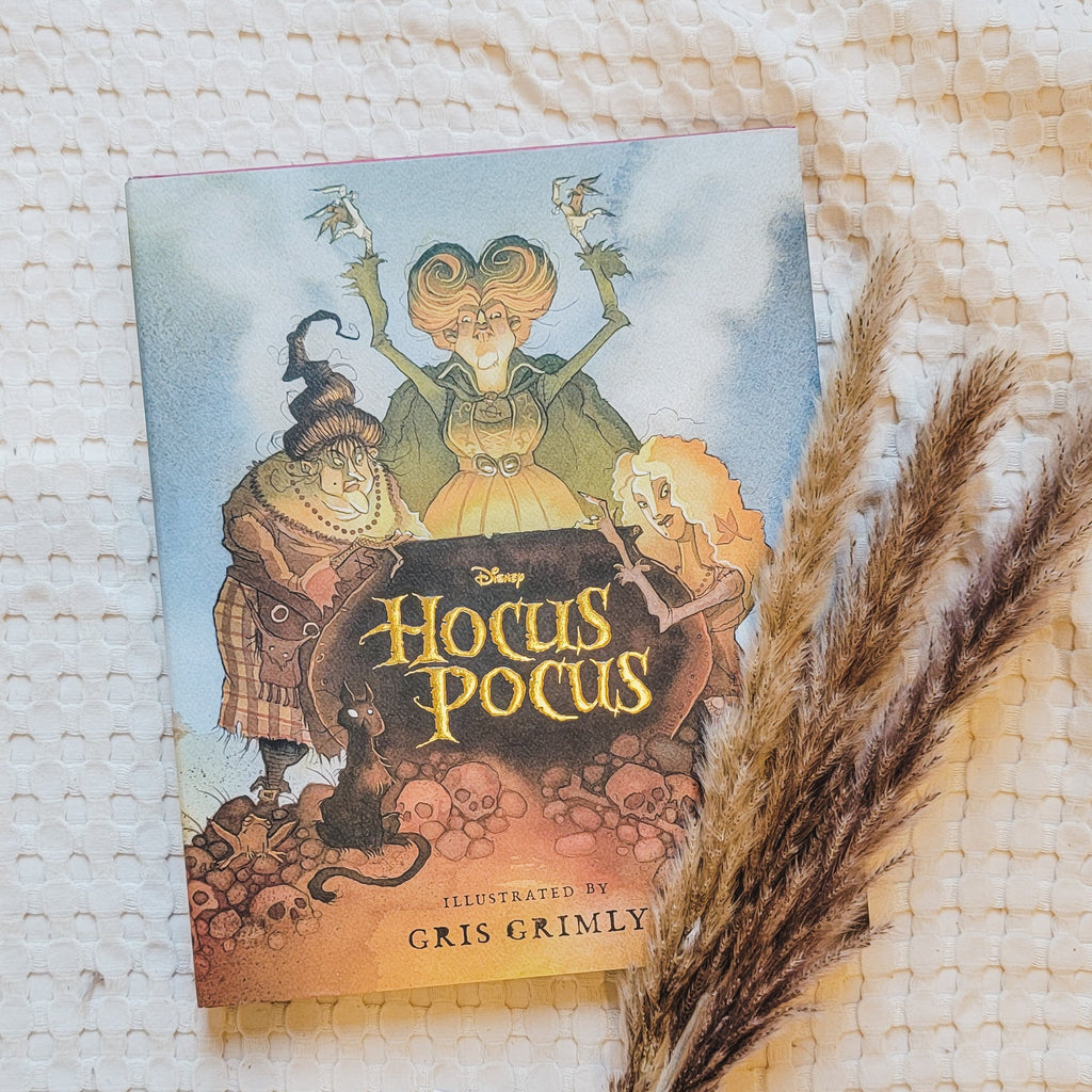Cream waffle blanket with pampas grass on the bottom right side, and the book Hocus Pocus: The Illustrated Novelization by A.W. Jantha. Cover shows the 3 sisters leaning over a black cauldron.