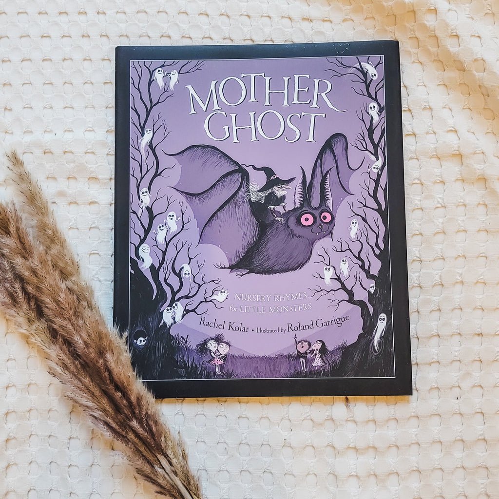 Cream waffle blanket with pampas grass on the bottom left corner, and the book Mother Ghost: Nursery Rhymes For Little Monsters by Rachel Kolar. Cover of the book is black and purple with white ghosts all over, and a large bat with a witch riding on his back.