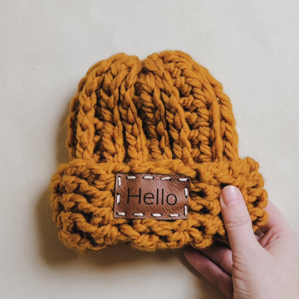 Cream background with the Handknit "Take Me Home" Beanie in Ochre/Hello by Petit Nordique. Beanie is a beautiful ochre colour with a hand sewn leather tag that says "hello".