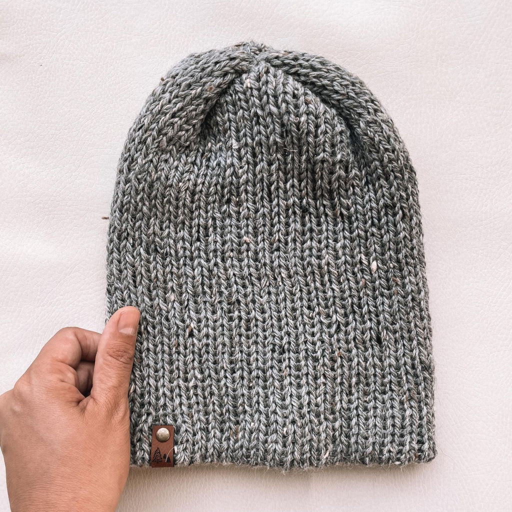 White background with Handknit Beanie by Petit Nordique in grey Tweed, and a hand holding on the side. Grey Tweed yarn, with a small leather tag.