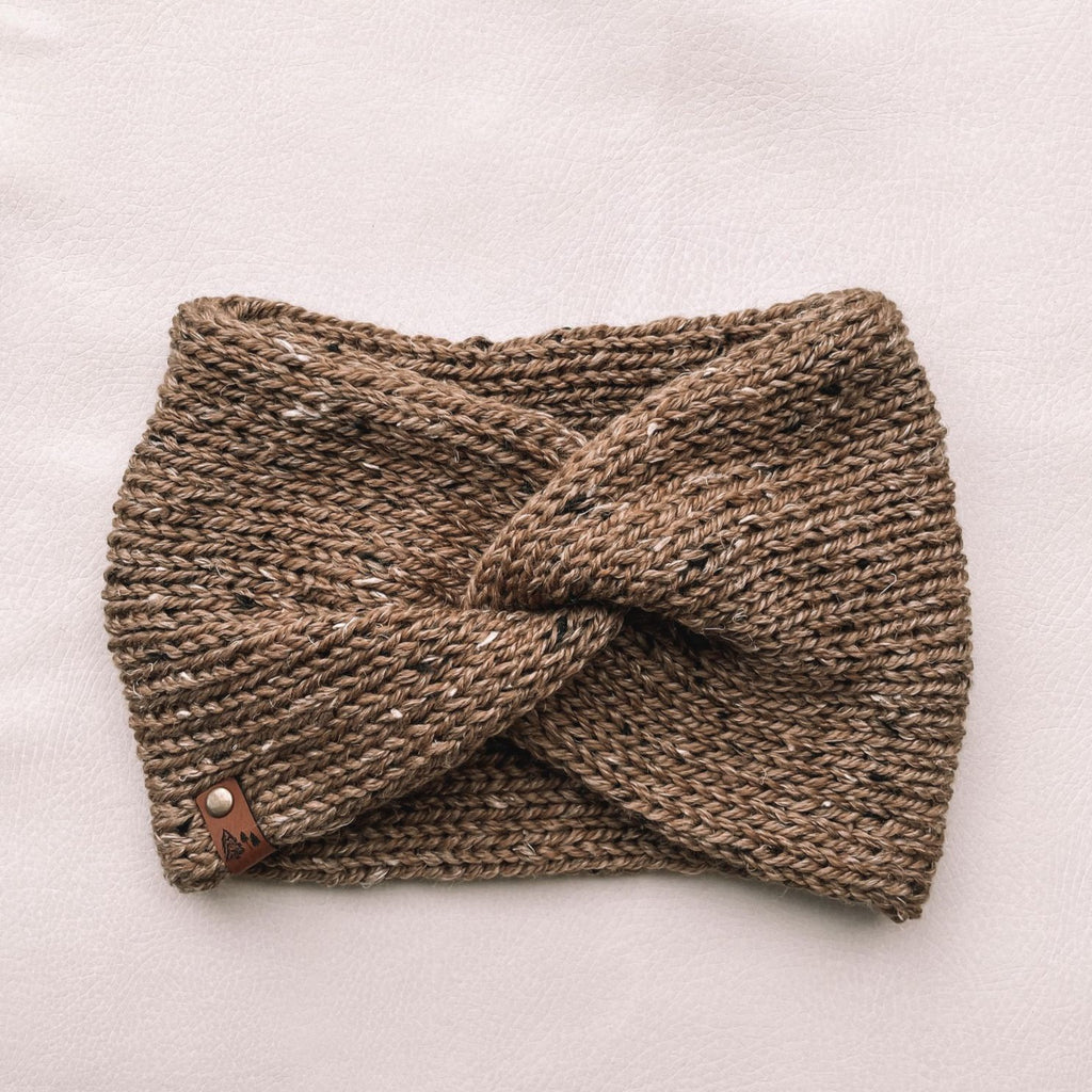 White background with Handknit Twist Headband by Petit Nordique in Ochre Tweed. Headband has a twist front with a small leather tag, in an ochre tweed yarn.