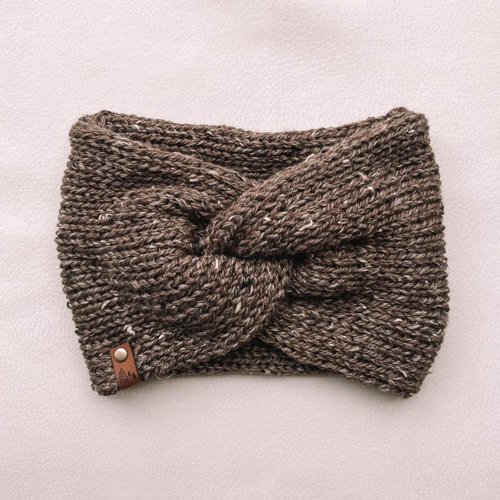 White background with Handknit Twist Headband by Petit Nordique in Brown Tweed. Headband has a twist front with a small leather tag, in a brown tweed yarn.