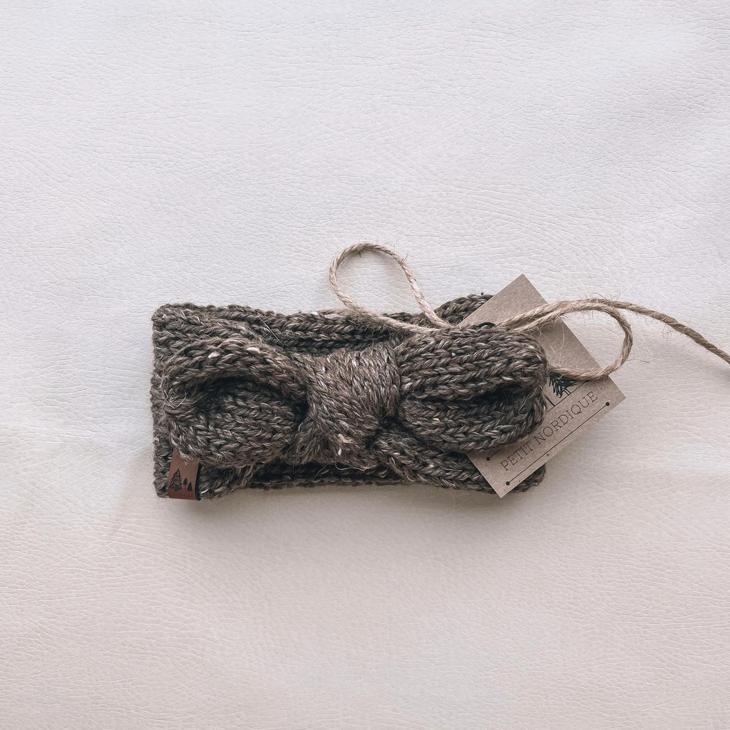 White background with Handknit Knot Bow Headband by Petit Nordique in Brown Tweed. Headband has a knot in the front with a small leather tag, in a brown tweed yarn.
